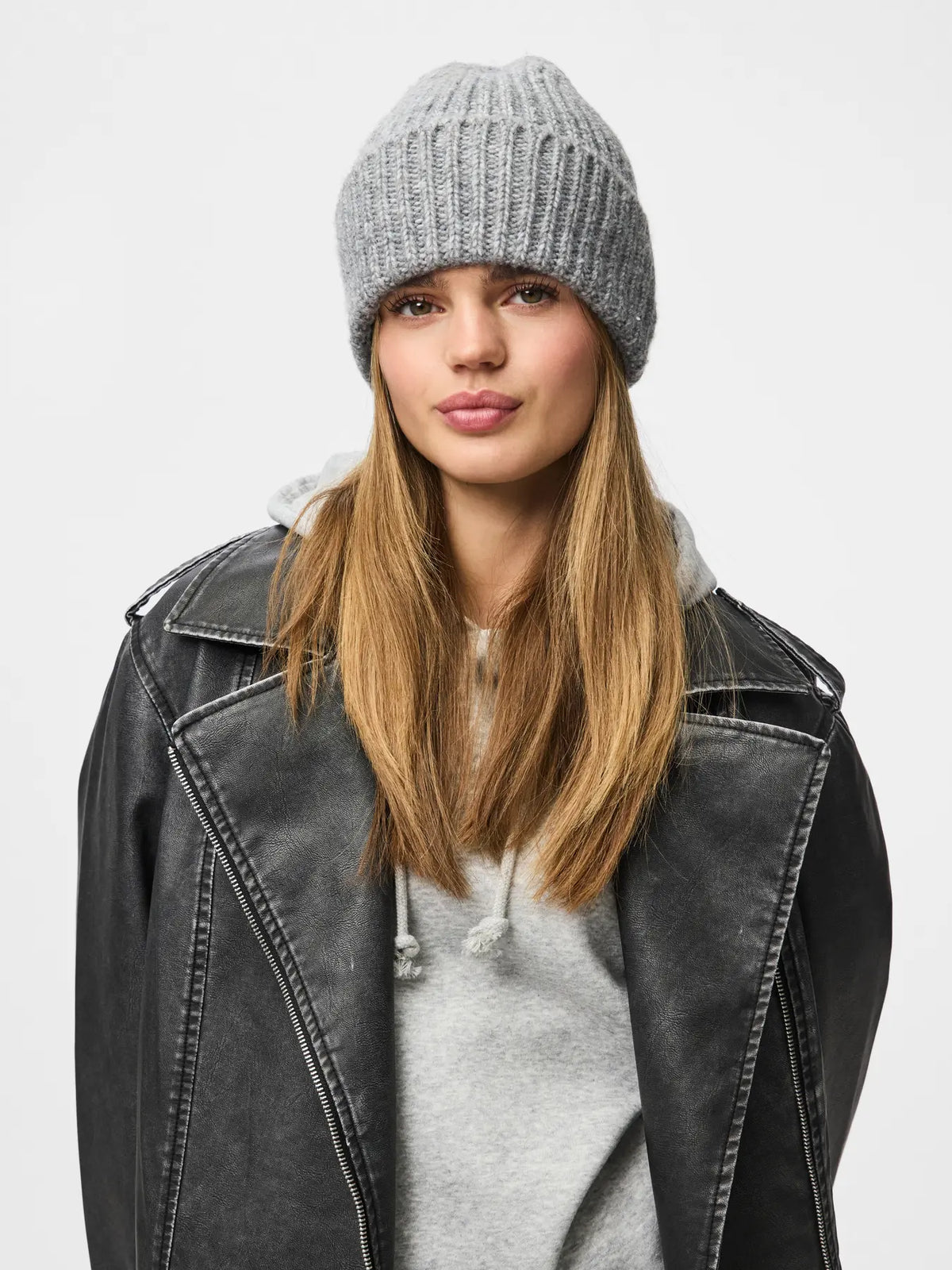 A woman wearing a light gray ribbed knit beanie, a gray hoodie, and a black leather jacket, photographed against a plain light gray studio background.