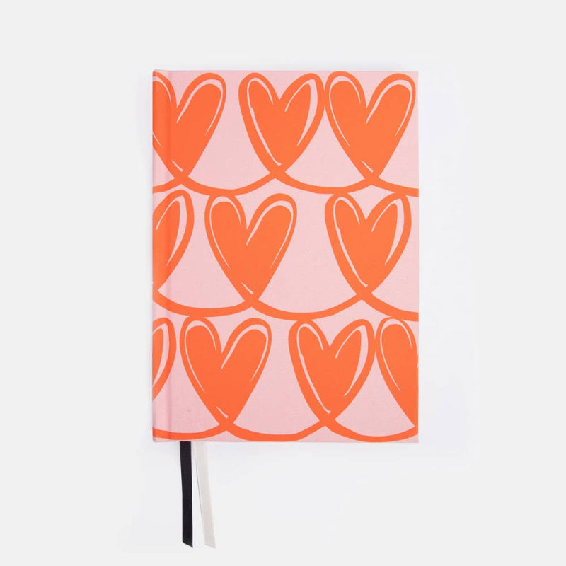 A hardcover notebook with a pink cover decorated with a repeating pattern of orange hearts, shown against a plain white background with a black and white ribbon bookmark.