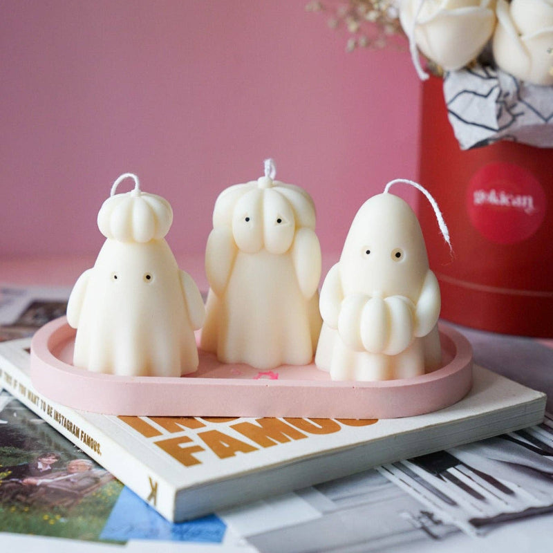 Three ghost-shaped candles on a pink tray with a magazine and flowers in the background.