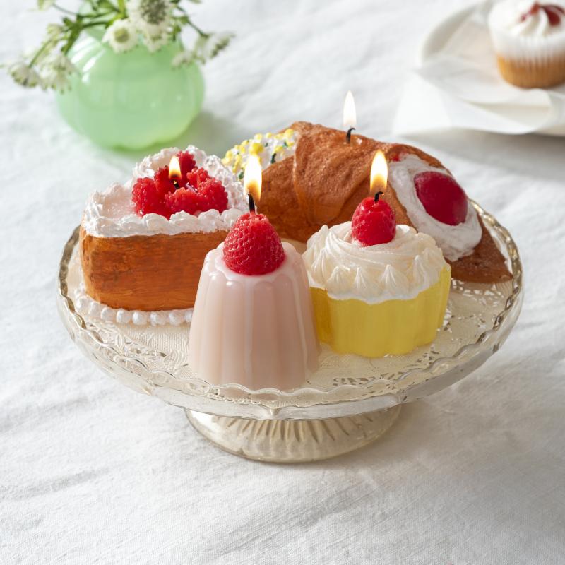 Decorative candles shaped like pastries and fruits on a white surface with a vase in the background.
