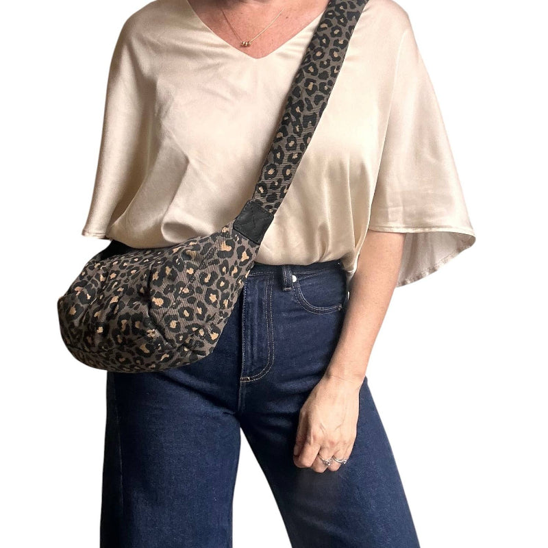 A woman wearing a cream satin blouse and dark blue jeans carries a leopard-print crossbody bag with a wide strap.