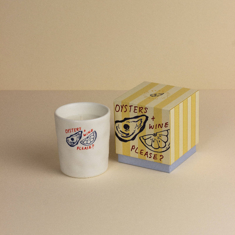 White candle with 'Oysters + Wine Please?' design next to a matching box on a beige background