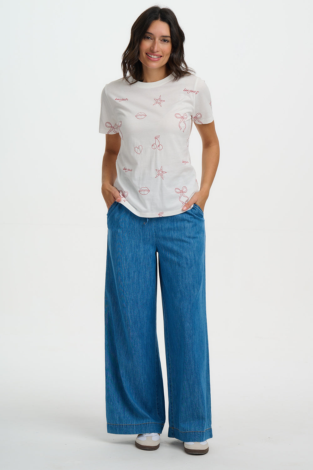 Woman wearing a Sugarhill Maggie T-Shirt with red patterns and blue pants on a white background