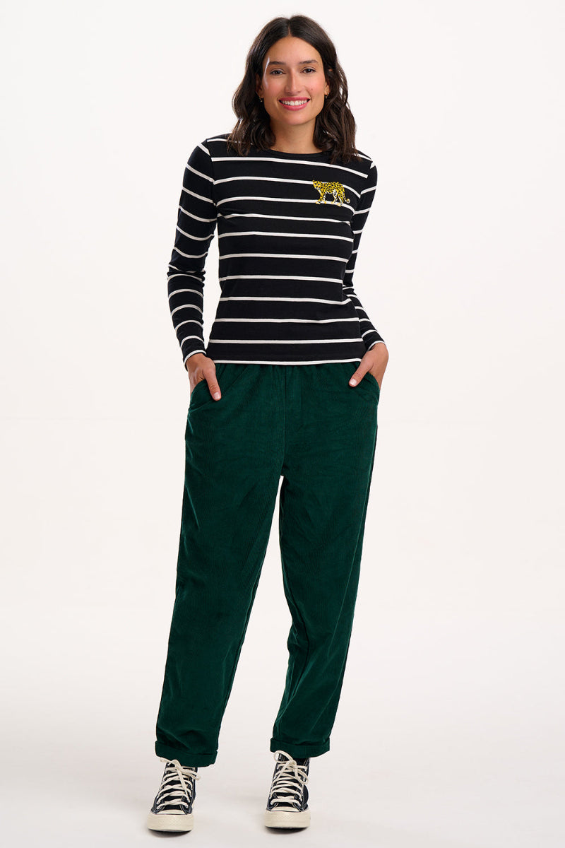 Woman wearing Sugarhill Vonnie Trouser Green a black and white striped sweater with a yellow logo and green pants on a white background