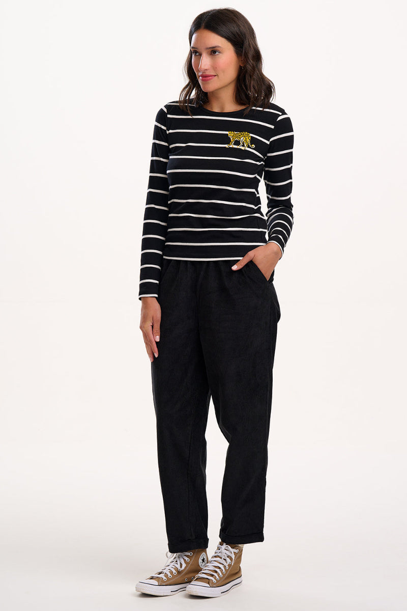 Woman wearing Sugarhill Vonnie Trouser a black and white striped sweater with a yellow logo, black pants, and brown sneakers on a white background.