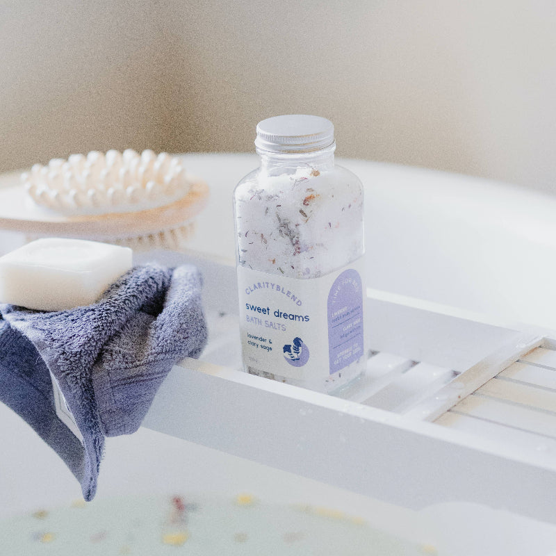 A clear plastic bottle of ClarityLabs Sweet Dreams Bath Salts sits on a white bathtub tray, surrounded by bath accessories including a bar of soap on a blue towel, a wooden brush, and a white tub.