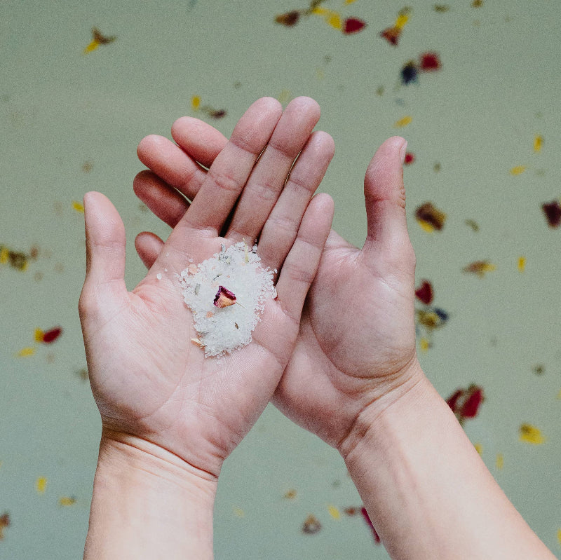 Two hands cupping a small pile of white flower petals with a single red petal, set against a soft-focus background of floating colorful petals.