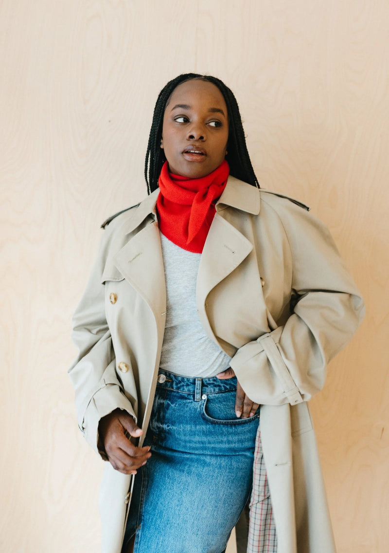 Woman wearing a beige trench coat, red scarf, and blue jeans against a plain background