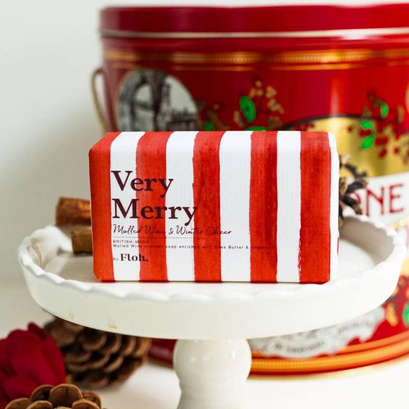 A wrapped gift box with red and white vertical stripes sits on a white pedestal. The front label reads 'Very Merry' and 'Melted Wool & Winter Solace' with the brand 'Floh.