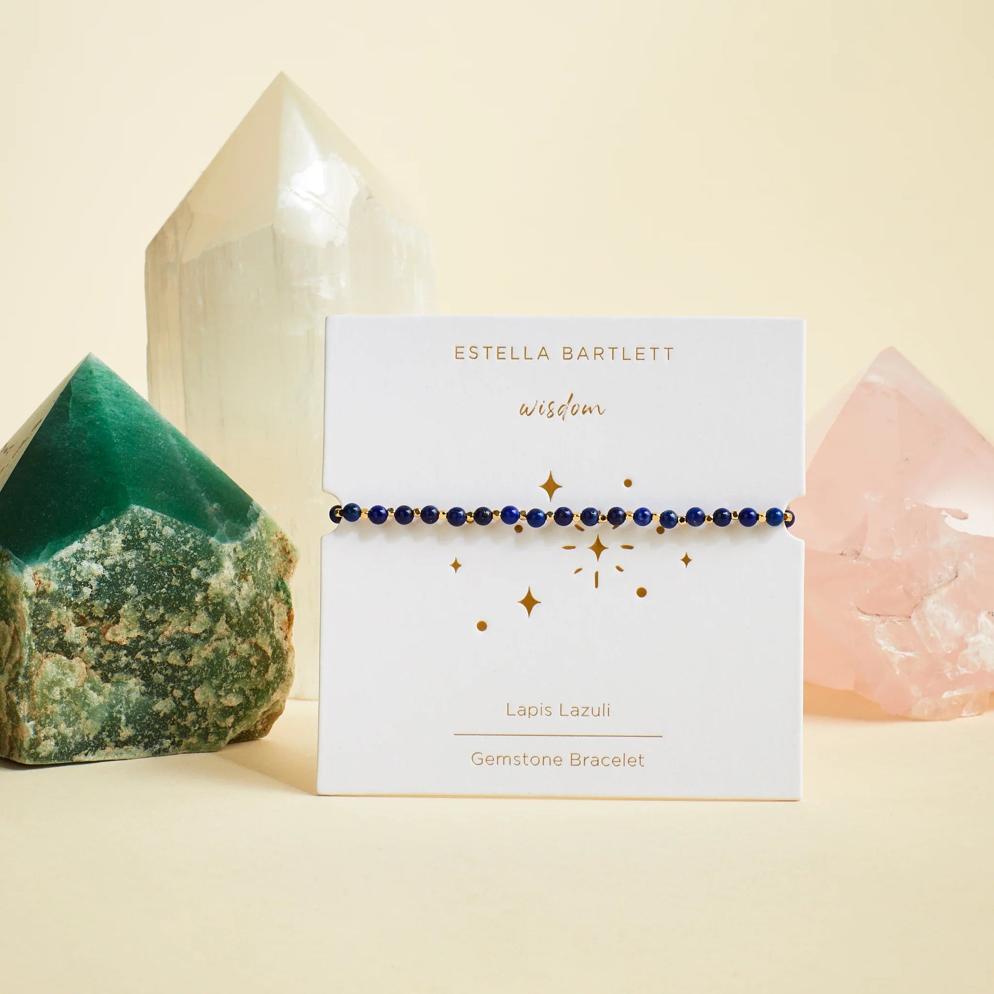 A delicate gemstone bracelet by Estella Bartlett, featuring deep blue round beads, displayed on its white branded card. The card is styled on a warm cream background, flanked by green, white, and pink crystal points.
