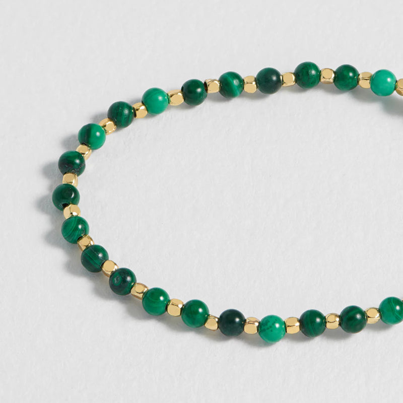 A beaded stretch bracelet featuring alternating round green beads and small gold spacer beads, displayed on a light gray textured background.