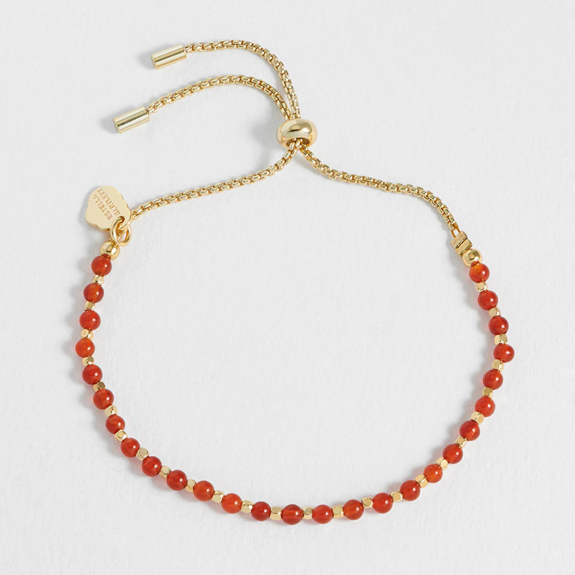 A delicate gold-tone adjustable chain bracelet with small, faceted orange-red beads, a small gold heart charm, and a gold logo tag, displayed on a clean white background.