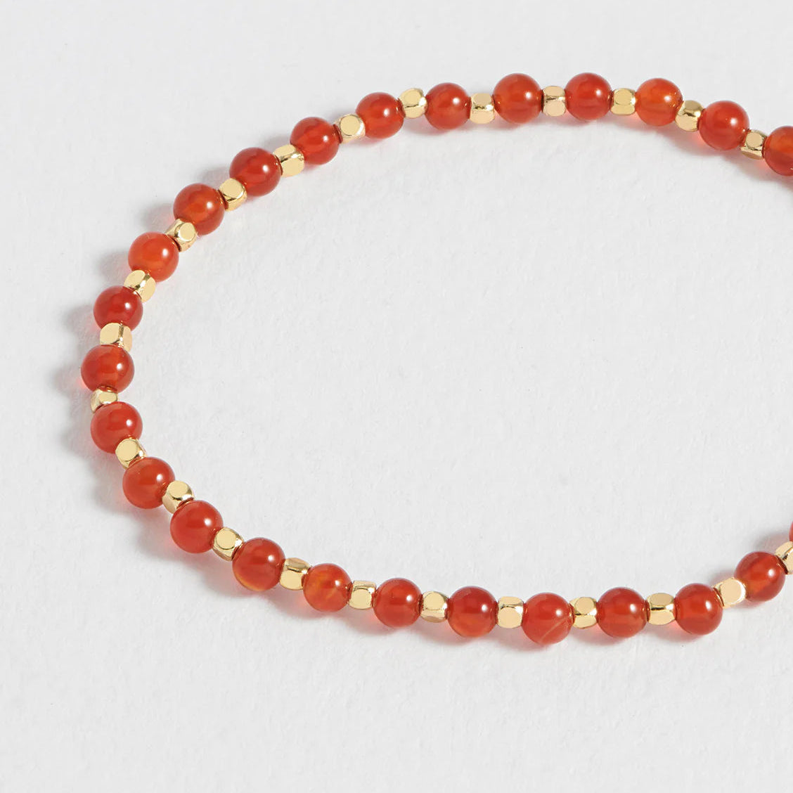 A beaded stretch bracelet featuring round orange-red stone beads separated by small metallic gold spacer beads, displayed on a light gray background.