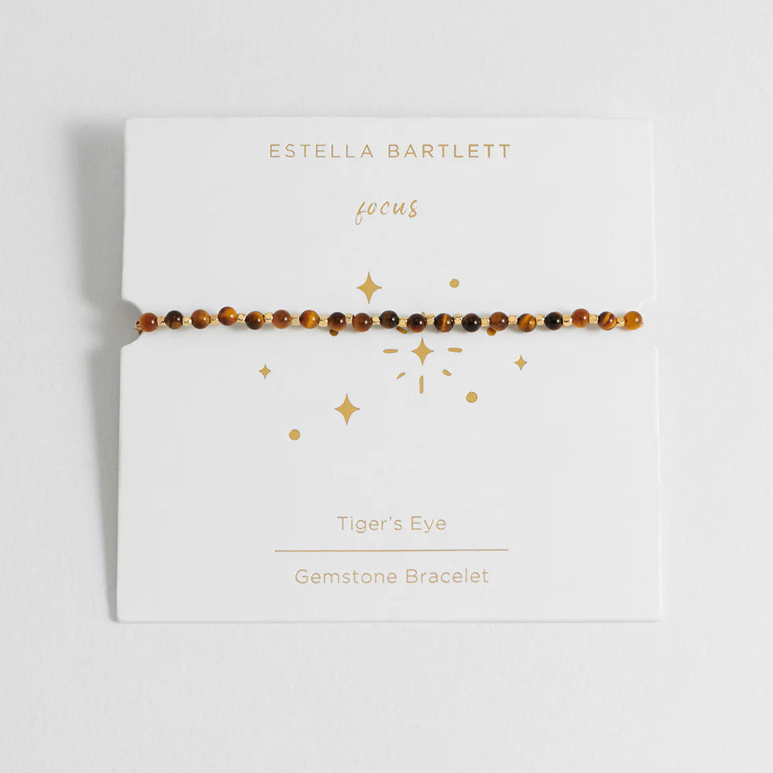 A tiger's eye gemstone bracelet by Estella Bartlett, displayed on its white card packaging with gold foil star illustrations.