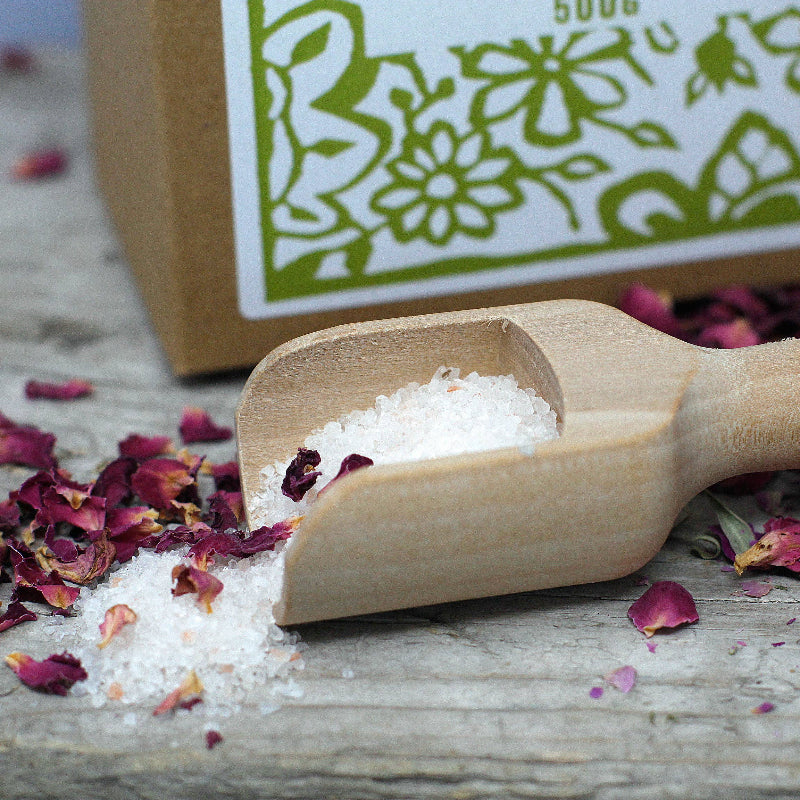 Wooden scoop filled with white powder on a wooden surface with rose petals and a branded box in the background.
