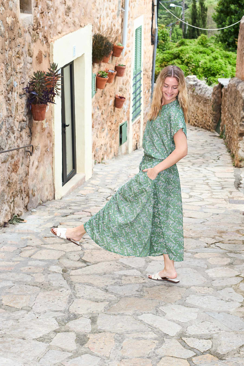 Woman in a green dress walking down a stone alleyway with plants and buildings.