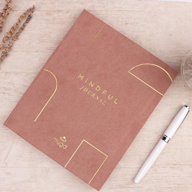 pink 'Mindful Journal' with gold text on a light wooden surface with a white pen.