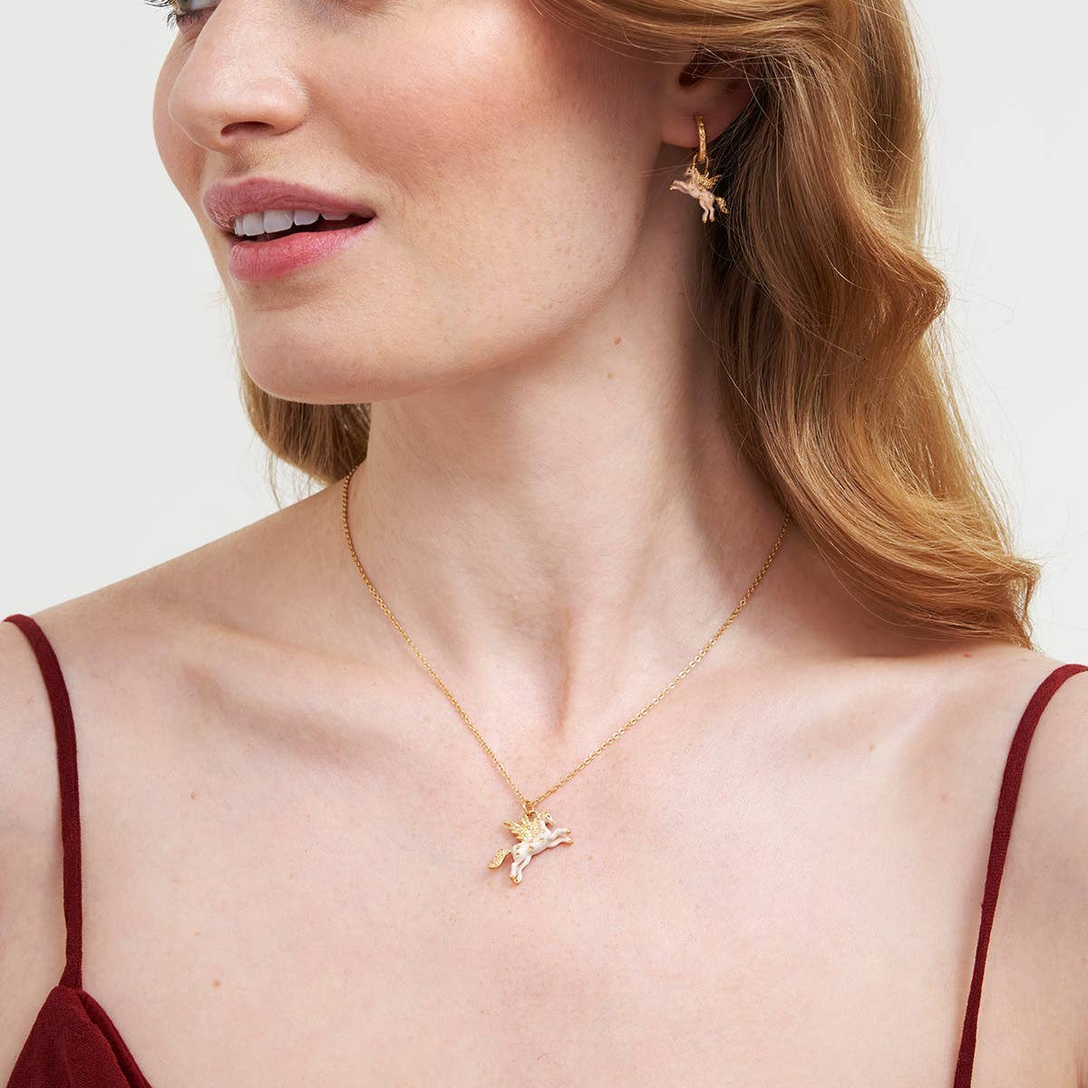 A close-up of a woman wearing a delicate gold necklace with a small starfish pendant and matching gold hoop earrings, paired with a burgundy camisole top against a light gray background.
