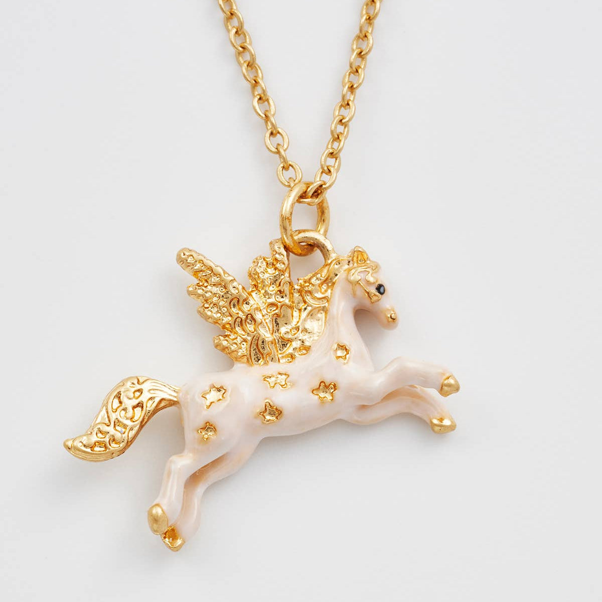 A gold-tone winged horse pendant necklace with a pale pink enamel body and gold star accents, shown on a matching chain against a light gray background.