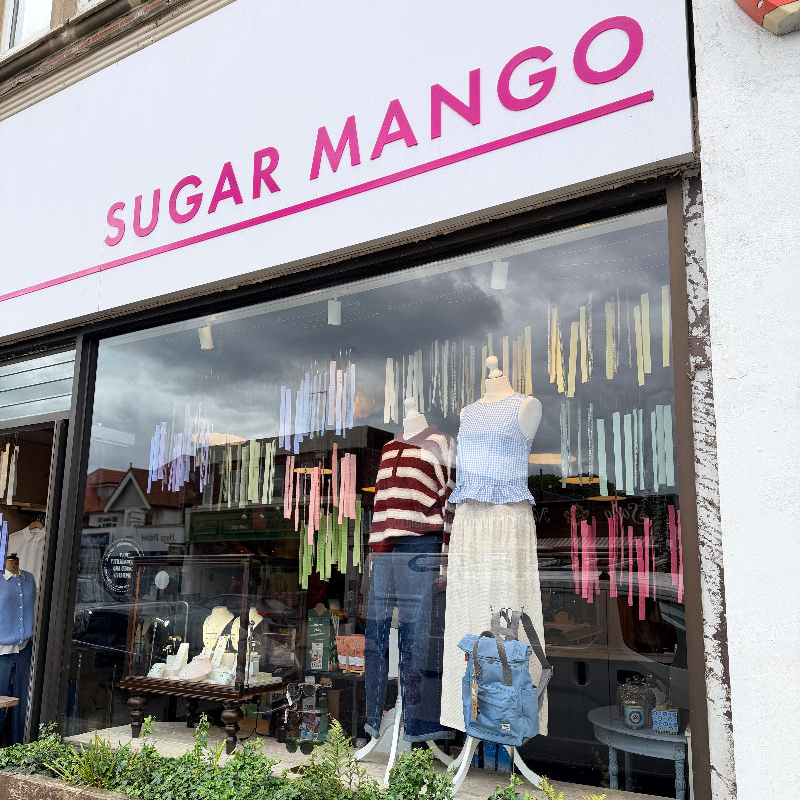 Sugar Mango clothing store storefront with a large window display featuring mannequins wearing a striped top and light blue skirt, with colorful hanging garments inside.