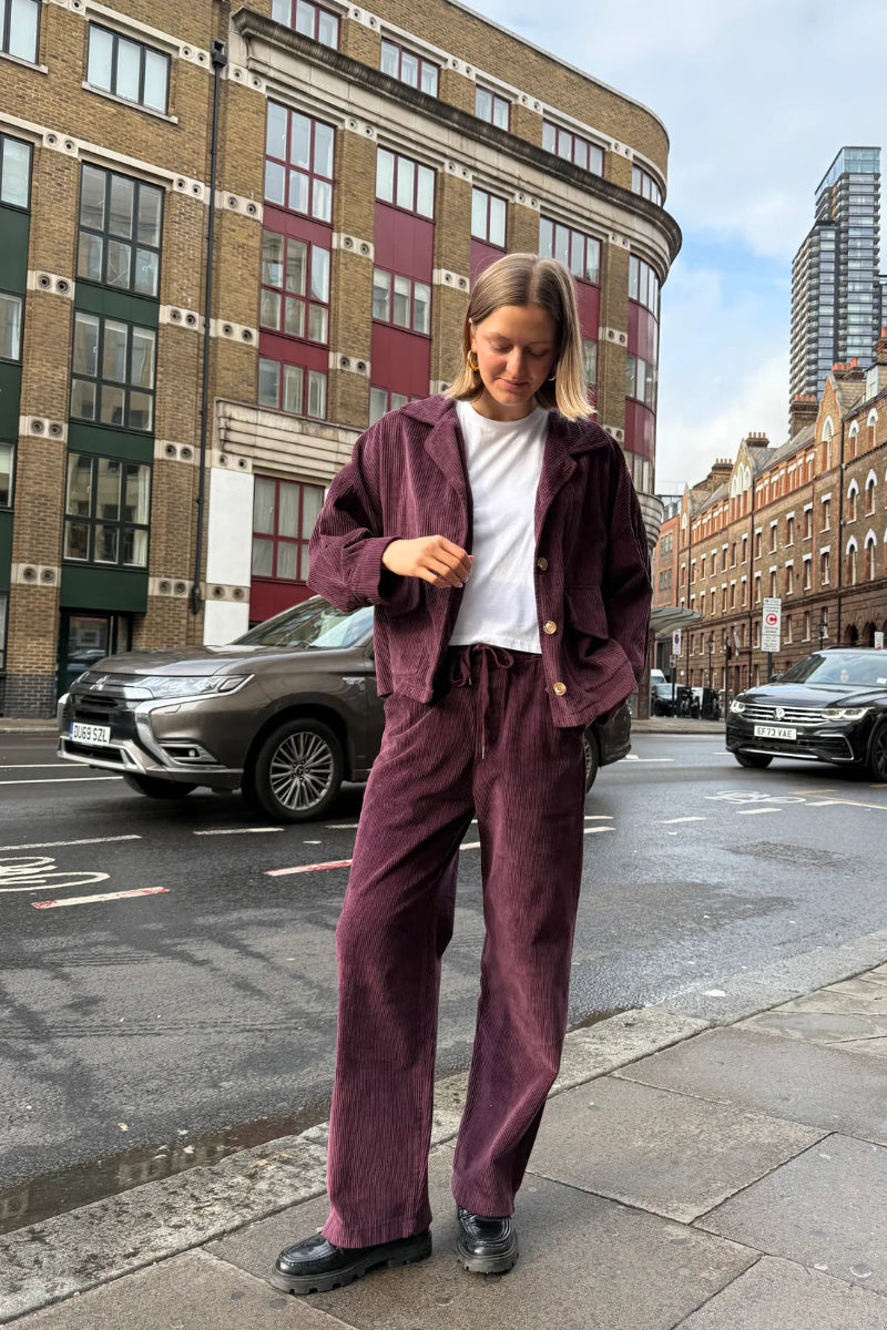 Person wearing a maroon tracksuit standing on a city street with buildings and cars in the background.