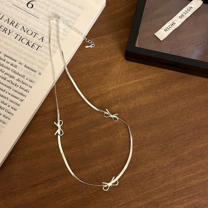 Silver necklace with bow details on a wooden surface next to an open book.