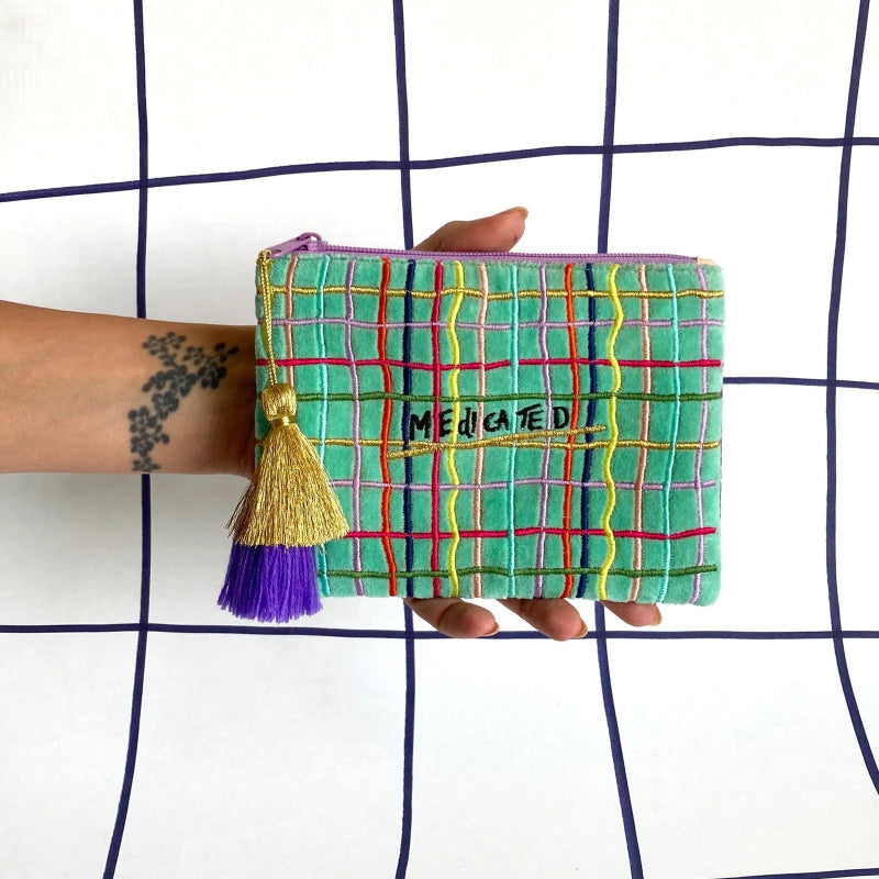 A hand holds a small zip pouch with a green plaid pattern, purple tassel, and the word 'MEDICATED' stitched on the front, set against a white background with a dark blue grid pattern.